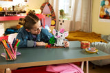 Child leaning across a table and touching the completed LEGO Disney Pua model, with a pink flower on Pua's head and a small hermit crab and green plant beside it.