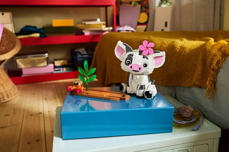 Completed LEGO Disney Pua model with a pink flower on its head, displayed beside a small hermit crab, green plant and wooden raft on a blue box in a furnished room.