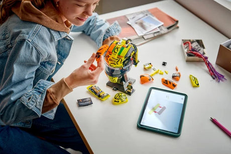 Person adding an orange piece to the LEGO Lando Norris Helmet at a desk with loose bricks, instruction booklet and digital building instructions on a tablet