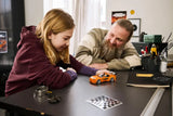 Two people at a table with a LEGO orange car model, sticker sheet and accessories