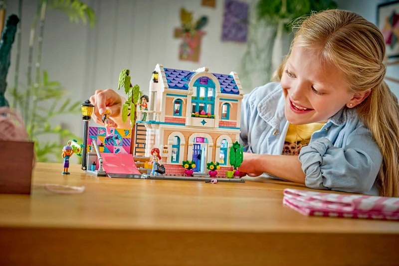 Child playing with LEGO Friends Liann's Family House 42687 on a wooden table, showing the completed house, colourful skate ramp, tree and minidoll figures.