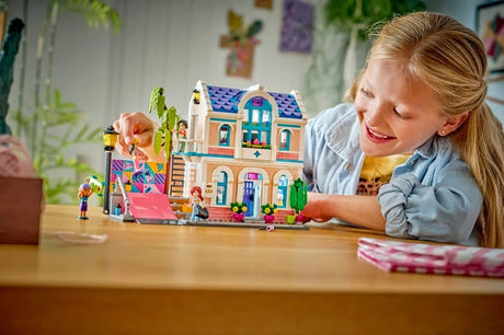 Child playing with LEGO Friends Liann's Family House 42687 on a wooden table, showing the completed house, colourful skate ramp, tree and minidoll figures.