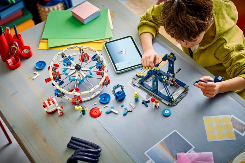 Child building a LEGO fairground model at a table with pieces, tools and instructions visible