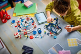 Child building a LEGO fairground model at a table with pieces, tools and instructions visible