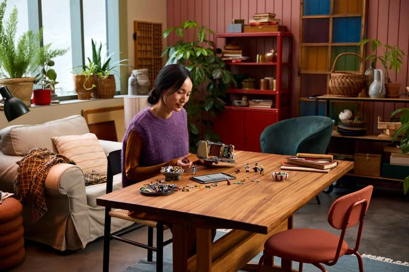 Adult seated at a table assembling LEGO pieces with minifigures and building sections laid out