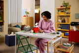 Woman assembling the brick built LEGO Snoopy's Doghouse at a table, with Snoopy and Woodstock on the red roof.