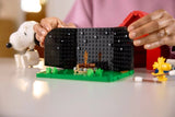 Hands opening the black hinged interior wall of the LEGO Snoopy's Doghouse scene, with a small campfire on the green base and Snoopy and Woodstock figures nearby.