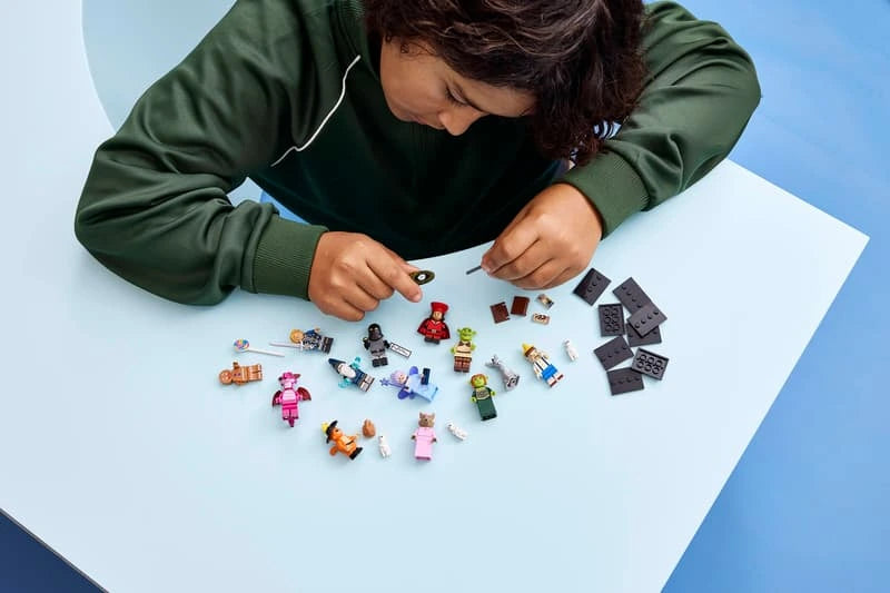 Person assembling LEGO Shrek minifigures at a table with characters, accessories and black display bases laid out