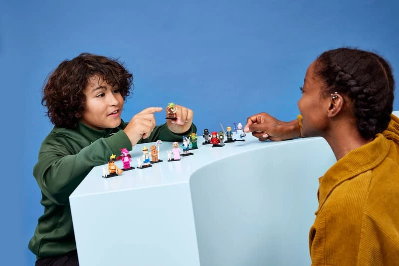 Two people holding LEGO Shrek characters across a row of assembled minifigures on a white display surface