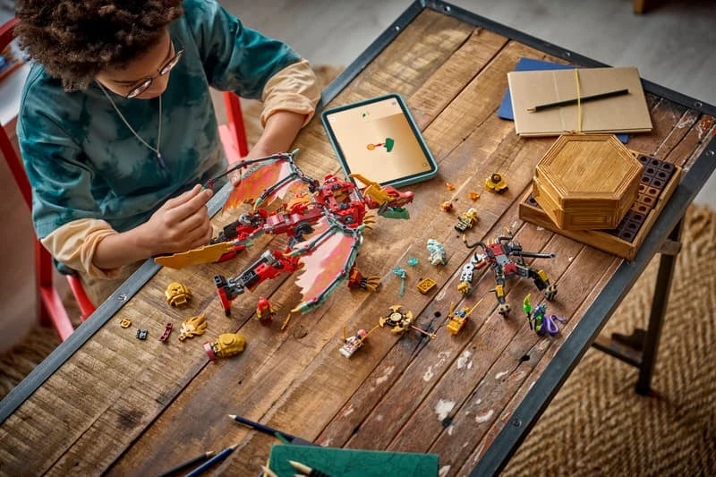Child building a LEGO NINJAGO dragon mech set on a table with pieces, minifigures and tablet instructions