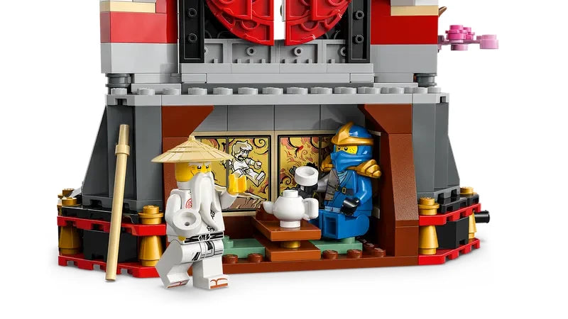 Rear view of the LEGO NINJAGO monastery display showing the open tearoom area with table, teapot, wall stickers depicting a battle scene, and two minifigures.

