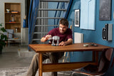 Man adjusting the completed LEGO Star Wars AT-AT walker on a wooden table, with the grey model and black display stand positioned in front of him.