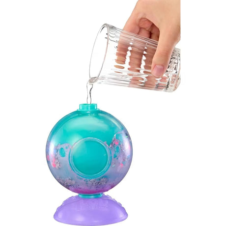 Hand pouring water from a glass into a transparent turquoise L.O.L. Surprise Bubbly Surprise capsule containing pink and purple fizz, mounted on a purple stand.