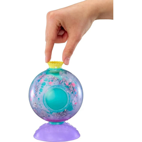 Hand placing a yellow cap onto the turquoise Bubbly Surprise capsule containing pink, blue and purple fizzy decorations, with a purple stand underneath.
