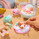 A hand tending to a Miniverse Real Baby Nurture miniature baby lying on a patterned changing mat over a pink cushion, with a second baby resting in a pale mint bassinet and miniature animal face baskets, nappies and toiletries nearby.