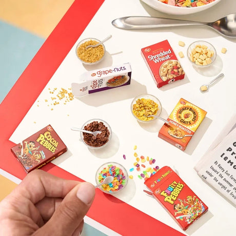 Overhead view of Miniverse miniature cereal boxes and bowls containing Fruity Pebbles, Cocoa Pebbles, Grape-Nuts, Shredded Wheat and Honey Bunches of Oats, with a hand holding a miniature spoon.