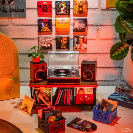Miniverse Real Music miniature record player with two speakers on a wooden stand, surrounded by album covers, vinyl records and storage crates, with more miniature album artwork displayed on the wall. Record player sold separately.