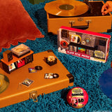 Miniverse Real Music promotional scene showing a boxed miniature record player, tiny vinyl records and album sleeves arranged on a suitcase beside a pink Real Music surprise capsule. Record player sold separately.