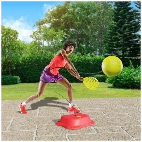 Person holding a yellow paddle beside the red Mookie Reflex Tennis Trainer base and yellow ball on a patio