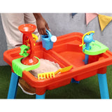 Child using a watering can at the Multiplay Table With Toys with sand in one side of the table