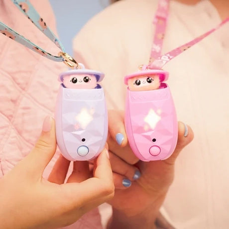 Two hands holding a lilac and a pink Peekimo interactive pet, both peeking out of their open pods with glowing star shaped lights and patterned neck straps.
