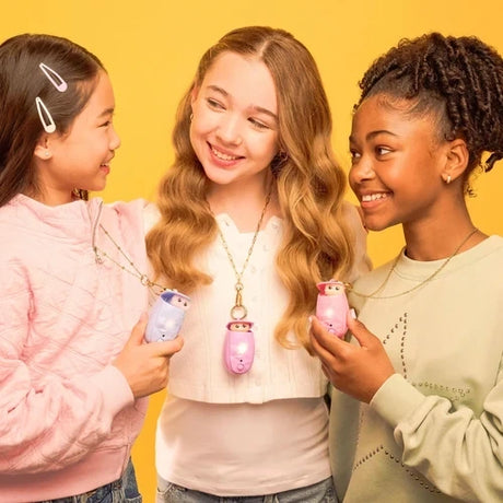 Three smiling girls against a yellow background, each holding or wearing a Peekimo interactive digital pet in pink or lilac, attached to a neck strap.