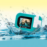 Rear view of the turquoise Photo Creator Waterproof Digital Camera surrounded by splashing water, showing its colour screen displaying two smiling children underwater and white camera control buttons.