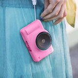 Close-up of the pink Photo Creator digital camera hanging from a white strap against turquoise clothing, showing the front camera lens and pink body.