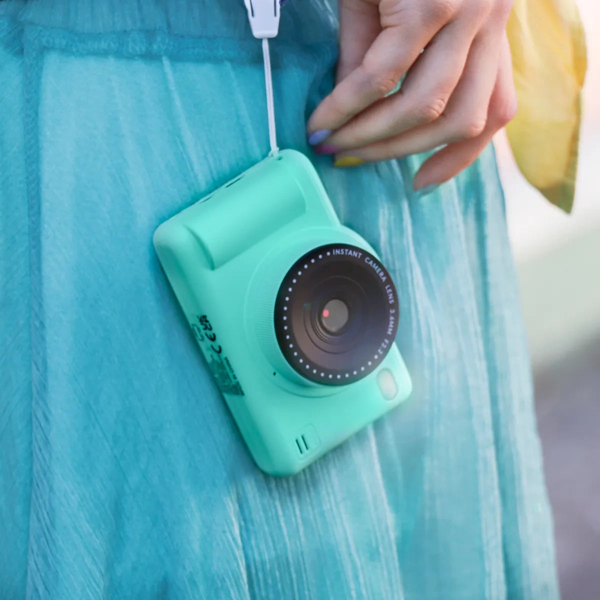 Green Photo Creator Pop Colour camera hanging by its strap against a turquoise dress, showing the camera's front lens, flash and compact body.