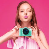 Woman holding the green Photo Creator Pop Colour camera at chest height against a bright pink background, wearing a pink top and posing with pursed lips.