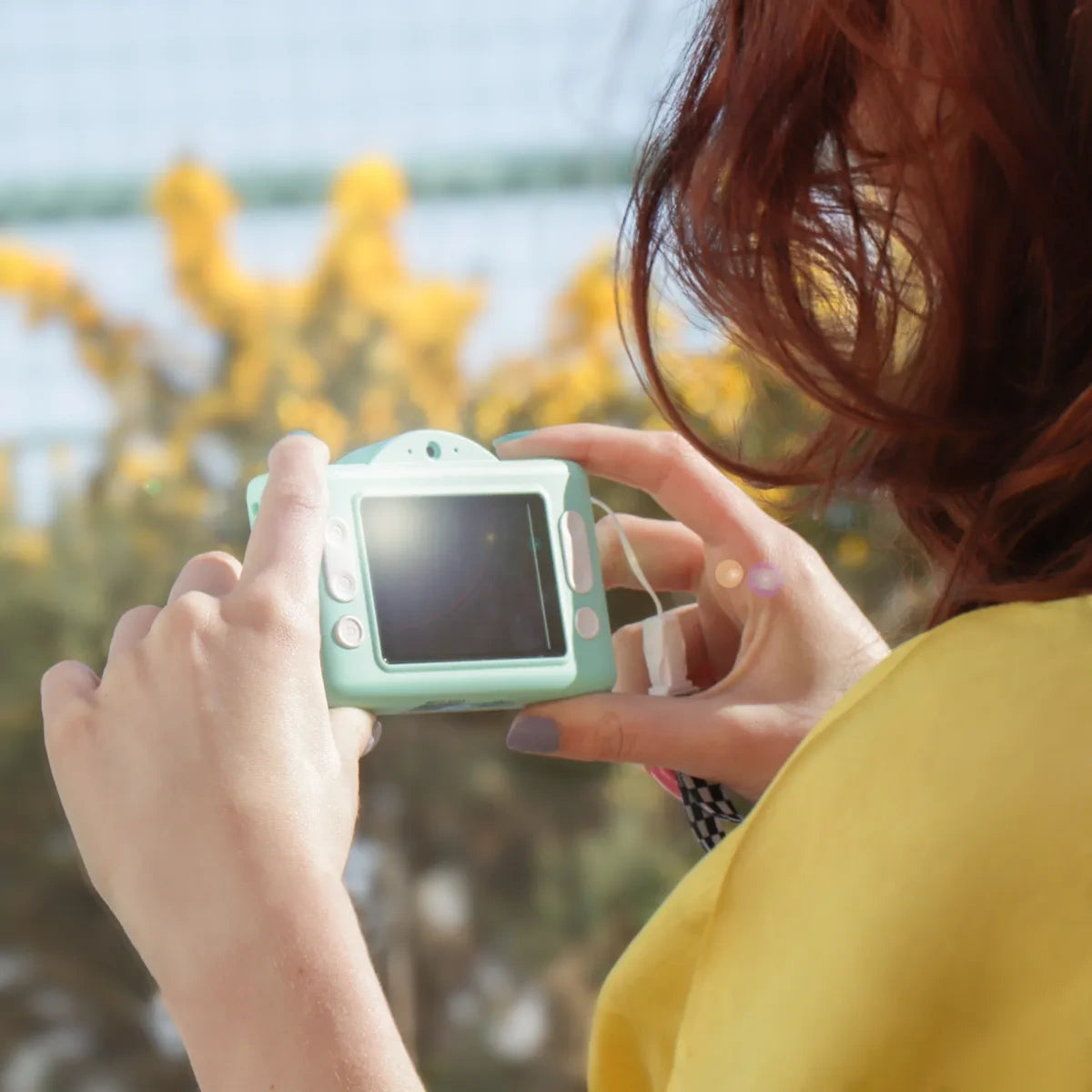Close-up from behind of a person holding the green Photo Creator Pop Colour camera outdoors, showing its rear screen, white control buttons and attached strap.