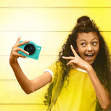 Girl in a yellow T-shirt holding the green Photo Creator Pop Colour camera at arm's length for a selfie, making a peace sign against a yellow wooden wall.