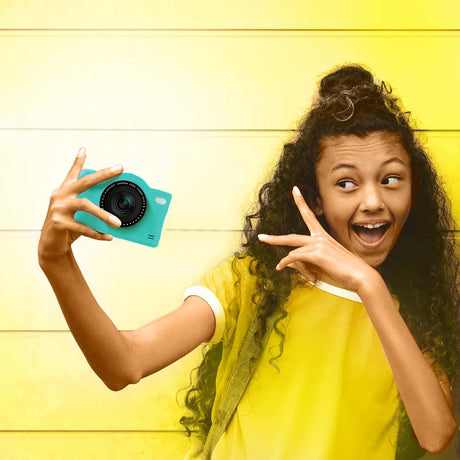 Girl in a yellow T-shirt holding the green Photo Creator Pop Colour camera at arm's length for a selfie, making a peace sign against a yellow wooden wall.