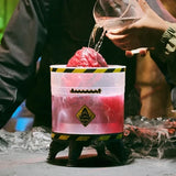 Hands pouring water from a clear jug onto the red Megalodon egg inside the Primal Hatch transparent hatching tank, with pink liquid, yellow hazard stripes and a shark warning symbol.