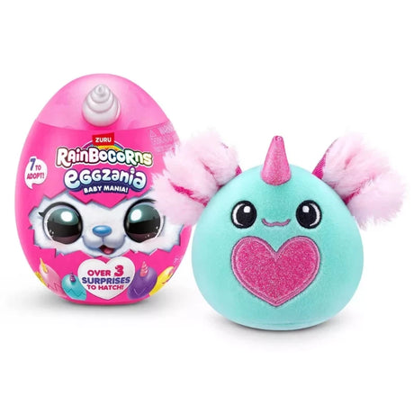 Teal Rainbocorns Eggzania Baby Mania plush with pink glitter heart and fluffy ears beside pink surprise egg showing “Over 3 Surprises To Hatch” text