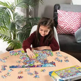 Child assembling the Ravensburger A Day in Chaosville jigsaw puzzle on a table with loose pieces surrounding the colourful town scene