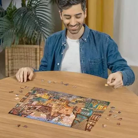 Man assembling the Ravensburger Bavaria Illuminated jigsaw puzzle on a table with loose pieces around the street scene