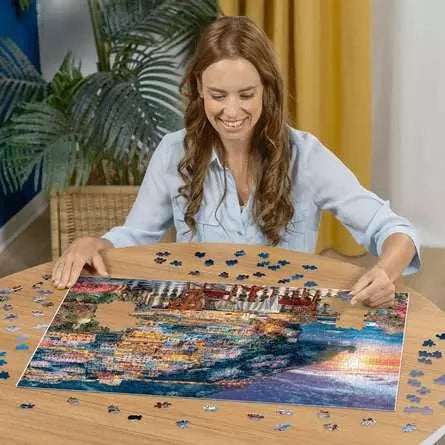 Woman assembling the Ravensburger Dinner in Positano jigsaw puzzle on a table with loose pieces around the colourful coastal scene