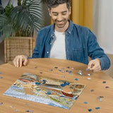 Man assembling the Ravensburger Happy Days at Work The Fisherman jigsaw puzzle on a table with loose pieces around the seaside scene