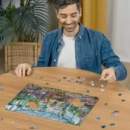 Man assembling the Ravensburger Morning in the Greenhouse jigsaw puzzle on a table with loose pieces around the greenhouse scene