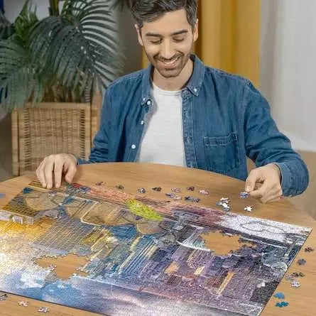 Man assembling the Ravensburger New York Summer and Winter jigsaw puzzle on a table with loose pieces around the image