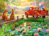 Two dogs beside a red pickup truck overlooking a lake with swans, lily pads and mountains