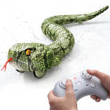 Close-up of the green amphibious RC snake with its long curved body, reptile scale pattern, yellow eyes and red tongue. Two hands operate a grey remote controller in the foreground, with water splashes surrounding the snake.
