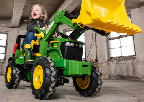 Child smiling while riding a Rolly Farmtrac John Deere 7930 ride on tractor toy with raised yellow loader in a bright indoor space