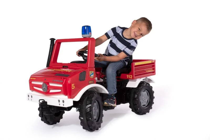 A child sitting on a red Rolly Fire Unimog pedal vehicle smiling while steering on a white studio background