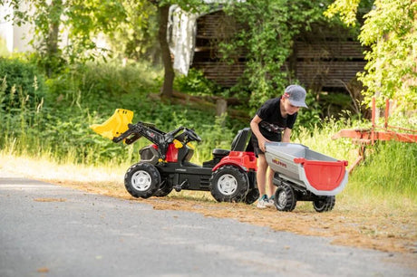 Child outdoors lifting the grey Rolly Halfpipe Single Trailer to empty its load beside a pedal tractor.