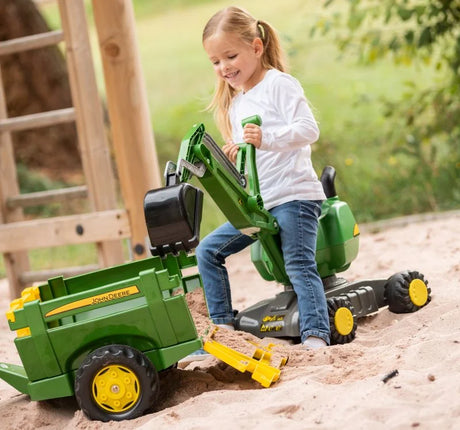 Child using the Rolly John Deere digger on wheels in a sandy play area while loading a green trailer