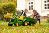Two children outdoors loading hay into a Rolly John Deere halfpipe trailer attached to a pedal tractor in a garden.