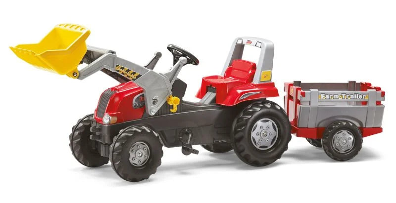Angled view of the Rolly Junior red tractor with loader raised and farm trailer attached on a white background