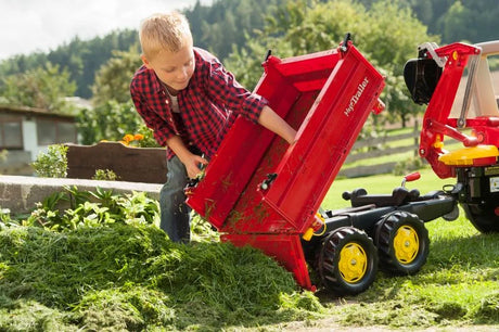 Child outdoors using the red Rolly Mega Trailer to tip a load of grass onto the ground during play.